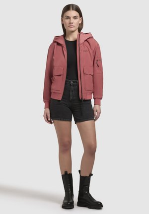 Giacca a vento rosa chiaro con cappuccio, tasche frontali, chiusura con zip e tasca a toppa sul maniche, abbinata a pantaloni corti neri a vita alta e stivali neri.