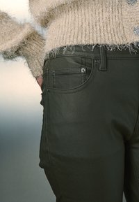 Maslinasto zelene traperice uskog kroja, s prednjim džepovima, metalnim kopčama i glatkom teksturom. Uparene s bež, mekanim džemperom.