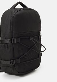 Sac à dos noir compact avec poche avant à cordon élastique, poche latérale en mesh, bretelles ajustables et poignée supérieure sur un fond clair uni.
