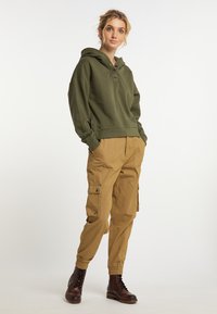 Felpa verde oliva con bottoni, fessure laterali, abbinata a pantaloni cargo color khaki con numerosi tacchi, e stivali marrone con lacci.