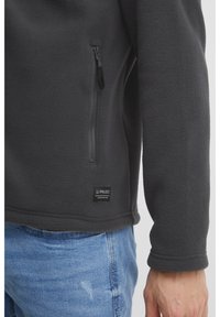 Flis pulover v temno sivi barvi, z zadrgo na žepu, teksturirano tkanino in oznako na spodnjem robu. Ohlapen kroj z zaobljenim ovratnikom.