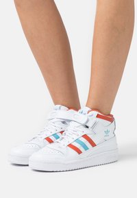 White high-top sneakers with red and blue stripes, featuring a strap, smooth leather material, and chunky sole. Accents include logo on heel.