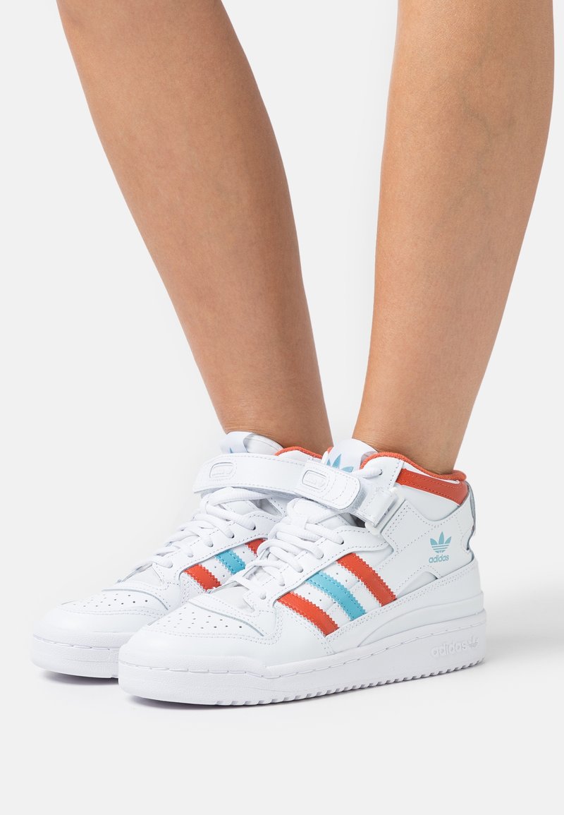 White high-top sneakers with red and blue stripes, featuring a strap, smooth leather material, and chunky sole. Accents include logo on heel.