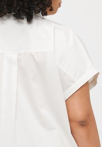 Chemise blanche à manches courtes avec un détail de pli au dos, présentant une texture de tissu lisse et des ourlets de manches retroussés.