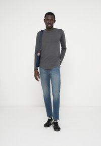 Grijze langemouwen shirt, blauwe spijkerbroek, zwarte sneakers met witte strepen en een blauwe schoudertas. Model staat tegen een witte achtergrond.