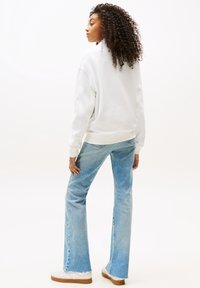 Sudadera blanca de corte relajado, combinada con unos vaqueros de tiro alto y pata de elefante de color azul claro y zapatillas blancas con suelas marrones. Cabello rizado visible.