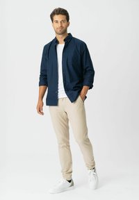 Man staat met één hand in zijn zak, draagt een marineblauw overhemd over een witte T-shirt, beige broek en witte sneakers op een witte achtergrond.