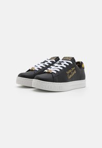 Baskets synthétiques noires avec lacets blancs, détails dorés et semelle blanche embossée avec le logo "COUTURE". Bout arrondi et texture lisse.