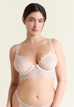 Frau trägt weißen, transparenten BH mit floralem Stickmuster und passende Unterwäsche, steht vor hellem, einfachem Hintergrund und schaut in die Kamera.