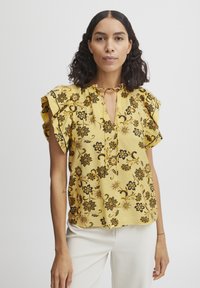 Blusa gialla floreale con fiori neri, maniche arricciate, scollatura a chiave e tessuto texturizzato. Abbinata a pantaloni chiari.