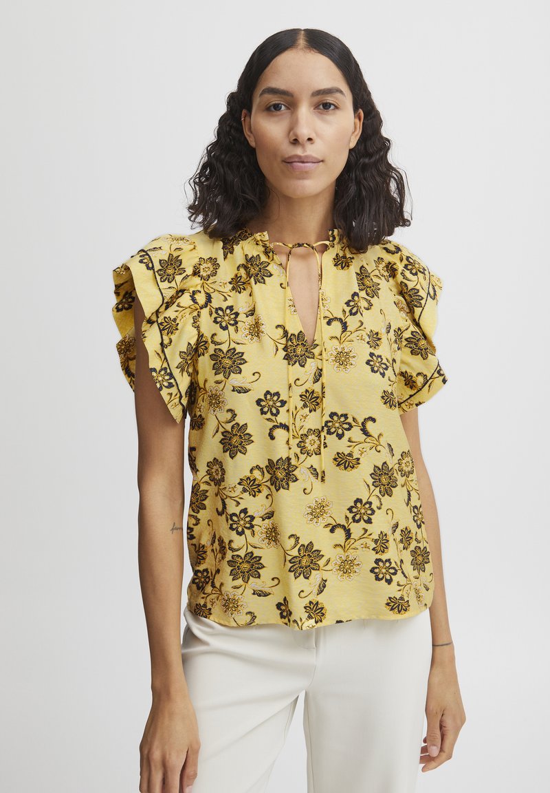 Blusa gialla floreale con fiori neri, maniche arricciate, scollatura a chiave e tessuto texturizzato. Abbinata a pantaloni chiari.