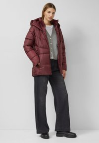 Bordeauxrode gewatteerde jas met capuchon en een glanzende textuur. Gecombineerd met een grijze cardigan en wijde zwarte denim broek. Zwarte laarzen maken de look compleet.