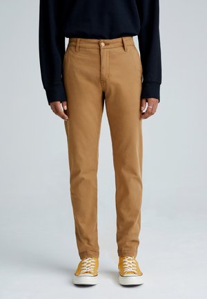 Osoba ubrana w beżowe spodnie o prostych nogawkach, żółte sneakersy sznurowane oraz czarną bluzkę z długim rękawem, stojąca na tle jasnego, gładkiego tła.