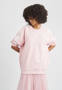 Ροζ φαρδιά φούτερ κατασκευασμένο από απαλό ύφασμα, με λευκό σχέδιο τριών ρίγων στα μανίκια και διακριτική λεπτομέρεια λογότυπου στο στήθος.