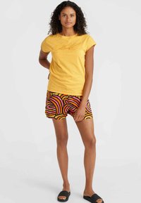 T-shirt gialla a maniche corte con stampa testurizzata, abbinata a pantaloni corti a fantasia nera, rosa e arancione. Infradito nere piatte.