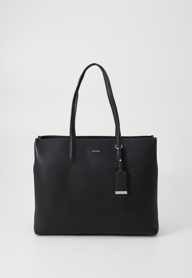 Black tote bag made of smooth synthetic material with two long handles, zip closure, and a detachable tag. Features a minimalist design.