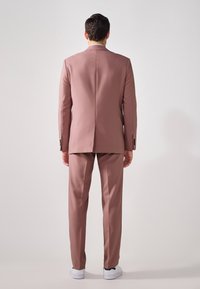 Helles rosa Anzug mit taillierter Jacke und passender Hose. Glattes Gewebe, einziger Schlitz hinten, getragen mit weißen Sneakers.