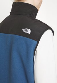 Blaues Fleece-Weste mit schwarzem Oberteil, hohe Kragen und weißem Logo. Strukturierter Stoff mit glatter Oberfläche an den Schultern.