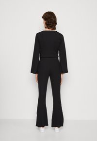 Person som står med ryggen mot kameran, iklädd en svart ribbad långärmad tröja och matchande utsvängda byxor, med vita sneakers, mot en vit bakgrund.