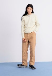 Chemise longue beige clair avec un logo triangulaire, pantalon beige, et chaussures de sport noires. Tissu doux et coupe décontractée.