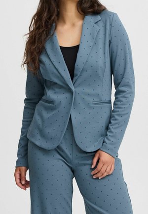 Žena oblečená v modrém saku a kalhotách se stejným vzorem malých tmavých puntíků, s černým topem pod tím, stojí s uvolněnýma rukama.