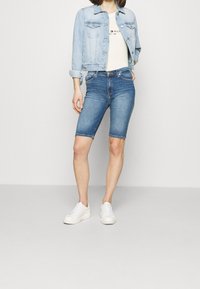 Džínové šortky světle modré barvy, po kolena, s hladkou texturou, v kombinaci s krátkou světle modrou džínovou bundou a bílými teniskami.