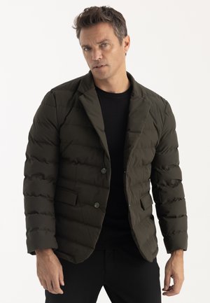 Homme portant un blazer matelassé vert olive foncé sur une chemise noire, debout devant un fond clair uni.