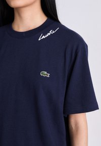 Nő, aki sötétkék pólót visel, amin egy kis zöld krokodil logó van a mellkasán, és a nyak körül a "Lacoste" felirat.