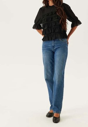 Kobieta ubrana w czarną bluzkę z falbanami, wysokie niebieskie dżinsy i czarne baleriny, stojąca z rękami za plecami.