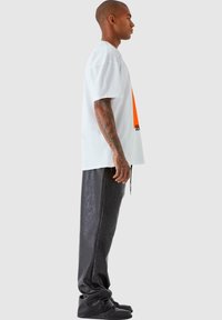 Hvit oversized t-skjorte med oransje grafikk og svart tekst, kombinert med svarte imitert skinnbukser og svarte sneakers. Synlige tatoveringer på armen.