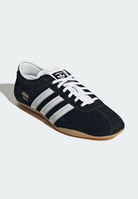 Svart mockaskor med vita randiga detaljer, rund tådesign, vita snörning och en beige gummisula; har "adidas"-logotyp.