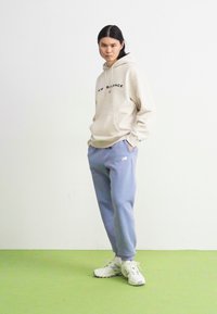 Hanorac bej cu text negru "New Balance", asortat cu pantaloni de trening albastru deschis și adidași albi. Croială casual și confortabilă.