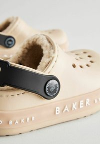 Zuecos beige hechos de material suave, con una correa negra y un forro interior de peluche. Texto blanco "BAKER by TED BAKER" en la parte inferior.