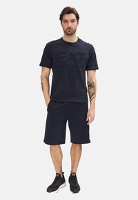 T-shirt in cotone blu navy con logo "EA7" in rilievo, abbinato a pantaloni corti blu navy, indossato con scarpe da ginnastica nere.