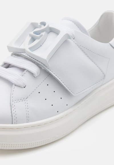 Vita lädersneakers med platt sula, som har en framträdande spänne och snörning, texturerad yta och små perforeringar för ventilation.