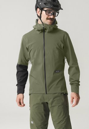 Mann trägt olivegrüne und schwarze Radjacke und Hose mit Helm und Schutzbrille und schaut nach unten.