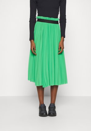 Woman wearing black long-sleeve top, bright green pleated midi skirt with ruffled waistband, and black athletic shoes standing against white background.