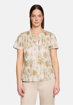 Vrouw met steil bruin haar, gekleed in een beige blouse met korte mouwen met een groene en bruine palmboomprint en beige broek, staand tegen een witte achtergrond.