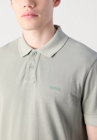Muž v světle šedém polo triku se dvěma knoflíky a logem "BOSS" na hrudi, zobrazený od krku po střed trupu na bílém pozadí.