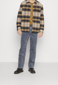 Kašmírová košile s kostkovaným vzorem v béžové, hnědé a námořnické modré barvě, s knoflíkovou přední částí a dvěma náprsními kapsami. V kombinaci s modrými kordovým kalhotami a černými botami.