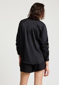 Person trägt ein locker sitzendes schwarzes Langarmshirt und schwarze Shorts, von hinten vor einem schlichten hellen Hintergrund gezeigt.