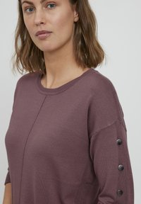 Lila kötött pulóver, kerek nyakkivágással, sima felülettel és díszes gombokkal az ujjakon a részletesség fokozása érdekében.