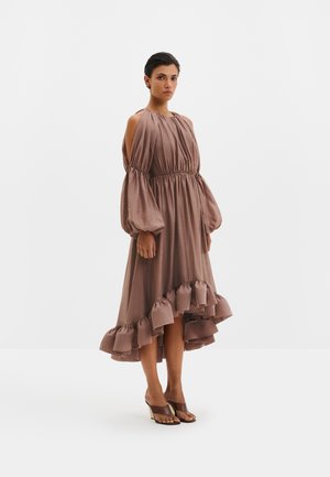 Femme debout portant une robe ample marron clair à épaules dénudées avec des manches ballon et un ourlet asymétrique à volants, assortie à des sandales à talons marron.