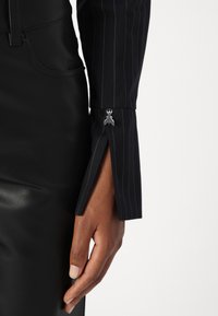 Tessuto a righe nere con un design elegante, caratterizzato da uno spacco laterale al polsino con un accent silver. Mano appoggiata su una superficie in pelle nera.
