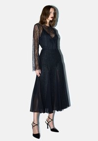 Top a maniche lunghe in pizzo nero abbinato a una gonna nera luccicante con pannelli trasparenti. Modella indossa tacchi a stiletto neri con cinturini.