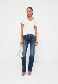 T-shirt bianca a scollo a V, jeans svasati in denim, tacchi grigi e una piccola borsa beige, in piedi di fronte a uno sfondo bianco.