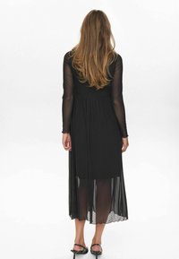 Rochie neagră cu mâneci lungi din tul cu buline, talie ajustată și fustă vaporasă din tul. Vederea din spate evidențiază textura și straturile.
