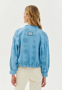 Světle modrá bomber bunda vyrobená z měkké tkaniny, zdobená vyšívanými květinovými vzory a nařaseným lemem. Obsahuje barevnou nášivku značky.