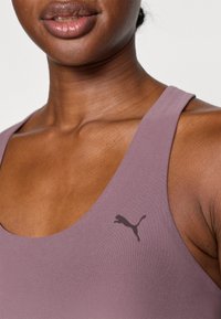 Biustonosz sportowy PUMA w jasnolawendowym kolorze, wykonany z gładkiego materiału, z dekoltem w kształcie litery U i małym czarnym logo po lewej stronie.