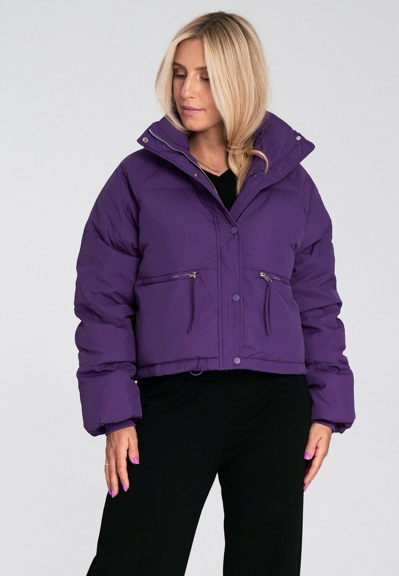 Krótka fioletowa kurtka puchowa z wysokim kołnierzem, zapinana na zamek błyskawiczny, z kieszeniami z przodu. Ma gładką teksturę, z zapięciami na guziki i elastycznymi mankietami.
